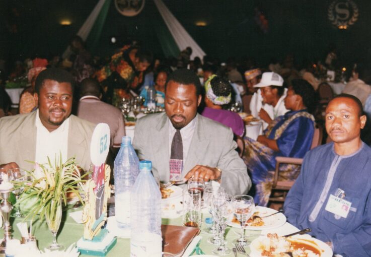 The African’s team, as part of a large African American delegation attending the inauguration of President-elect Olusegun Obasanjo, enjoys the lavish state dinner offered the night before the swearing-in. From left to right: photographer emeritus Arsene Kassegne; founder/publisher Soumanou Salifou; and reporter Aruna Adeyemi. Photographer unknown.