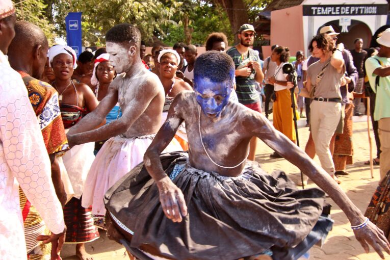 : Young male voodoo worshippers dancing in front of the pithing temple as foreign tourists watch, snapping pictures.